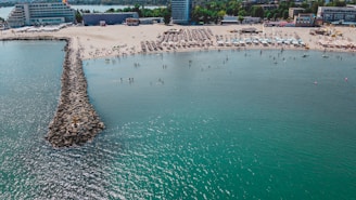 Aerial view of a beach with rows of sunbeds and umbrellas. The shoreline curves gently with a stone breakwater extending into the sea. People are scattered across the beach and swimming in the water. Buildings line the background, surrounded by greenery.