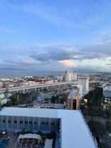 An urban landscape featuring several high-rise buildings, a busy highway with multiple lanes, and a sprawling cityscape extending to the coast. The sky is mostly clear with a few clouds, creating a calm and relaxed ambiance.