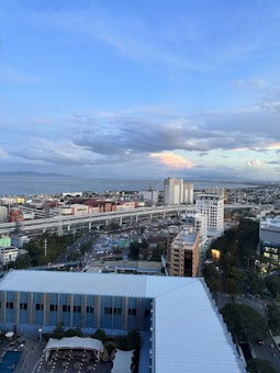An urban landscape featuring several high-rise buildings, a busy highway with multiple lanes, and a sprawling cityscape extending to the coast. The sky is mostly clear with a few clouds, creating a calm and relaxed ambiance.