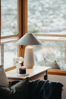 A cozy interior scene with a wooden side table placed beside a large window. On the table, there is a modern white lamp, a small bouquet in a white vase, and a scented candle on a coaster. Soft cushions and a blanket are arranged on the furniture nearby, adding to the comfortable atmosphere. The window reveals a blurred outdoor view, suggesting a serene environment.