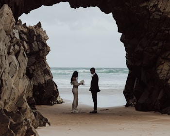 A couple stands facing each other at a sandy beach between large rocky cliffs, with the ocean waves in the background. The person on the left is wearing a long, flowing dress and appears to be holding a book or paper, while the person on the right is dressed in a suit.