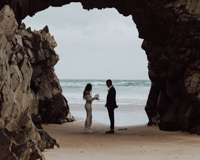 A couple stands facing each other at a sandy beach between large rocky cliffs, with the ocean waves in the background. The person on the left is wearing a long, flowing dress and appears to be holding a book or paper, while the person on the right is dressed in a suit.