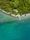 A solitary house with a gray roof nestled among lush green trees beside a tranquil turquoise lake. A wooden pier extends from the shore into the clear water, surrounded by a dense forest and a pebble beach.