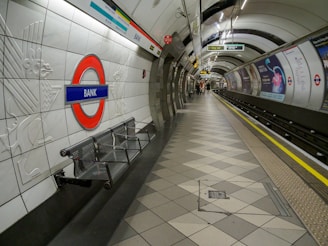 A London Underground station platform with the iconic roundel logo and the name 'Bank' displayed prominently. The platform is empty, with geometric-patterned flooring and metal benches. Advertisements line the curved walls, and signage indicates directions to different lines and exits. The lighting is bright, casting a clean, sterile atmosphere.