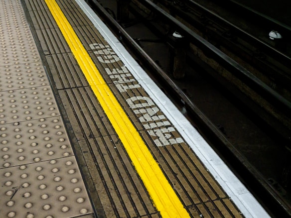 A subway platform edge with a tactile paving strip and a yellow safety line. The words 'MIND THE GAP' are painted on the edge near the tracks. The tracks are visible to the right.