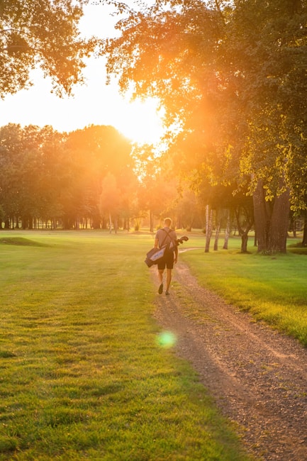 A person carrying a golf bag walks along a pathway through a green field surrounded by trees. The sun is setting, casting a warm glow over the scene.