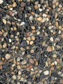 A close-up view of a gravel surface featuring a variety of small, smooth stones in different shades of brown, black, and gray. The stones are densely packed together, creating a colorful and textured pattern.