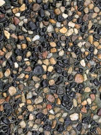 A close-up view of a gravel surface featuring a variety of small, smooth stones in different shades of brown, black, and gray. The stones are densely packed together, creating a colorful and textured pattern.