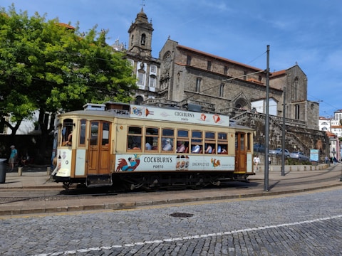 A vintage tram is traveling along a cobblestone street in a city. The tram is cream-colored with wooden accents and features advertisements on the side. Behind the tram, a historic stone building with arched windows and a bell tower is visible. The sky is blue, and there are lush green trees alongside the street.