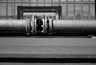 A black and white image featuring a metal pipe with a joint or valve at its center, positioned horizontally. The background consists of a series of vertical bars or railings.