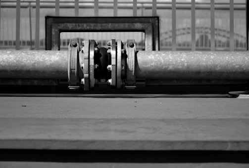 A black and white image featuring a metal pipe with a joint or valve at its center, positioned horizontally. The background consists of a series of vertical bars or railings.