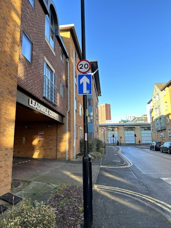 A street scene featuring a building with a brick facade, prominently displaying the name 'Leadmill Court.' A road sign indicates a speed limit of 20 and a one-way direction. The street is quiet, with some parked cars visible in the background. The sky is clear and blue, creating bright lighting with shadows cast on the building.