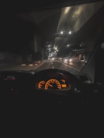A nighttime view from inside a vehicle, showcasing a brightly lit dashboard with orange speedometer and gauges. The road ahead is illuminated by streetlights, with faint reflections on the windshield. Several vehicles are visible in the distance, driving along a multi-lane road.