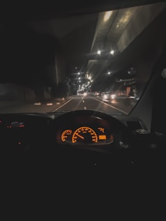 A nighttime view from inside a vehicle, showcasing a brightly lit dashboard with orange speedometer and gauges. The road ahead is illuminated by streetlights, with faint reflections on the windshield. Several vehicles are visible in the distance, driving along a multi-lane road.
