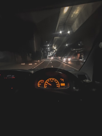 A nighttime view from inside a vehicle, showcasing a brightly lit dashboard with orange speedometer and gauges. The road ahead is illuminated by streetlights, with faint reflections on the windshield. Several vehicles are visible in the distance, driving along a multi-lane road.
