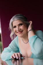 A mature woman with gray hair styled elegantly is smiling warmly. She is wearing a light teal blouse with a low neckline and jewelry including a necklace and watch. Her hand rests gently on a reflective surface, adding a serene feel to the portrait. The background is a rich, deep red color that contrasts softly with her attire.