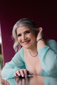 A mature woman with gray hair styled elegantly is smiling warmly. She is wearing a light teal blouse with a low neckline and jewelry including a necklace and watch. Her hand rests gently on a reflective surface, adding a serene feel to the portrait. The background is a rich, deep red color that contrasts softly with her attire.