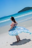 A person stands on a sandy beach, wearing a colorful swimsuit and a blue patterned beach wrap. In the background, the turquoise sea meets a clear sky, and a forested hill appears in the distance.