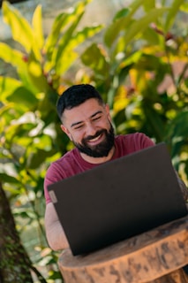 A person with a beard is sitting outdoors using a laptop, smiling and appearing engaged. Lush green plants fill the background, suggesting a garden or nature setting.