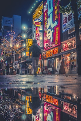 A vibrant urban street scene at night features brightly lit signs with Japanese characters. A large puddle on the ground creates a striking reflection of the illuminated buildings and signs, adding to the aesthetic appeal. A person wearing a hooded jacket walks along the street, capturing a moment of city life.