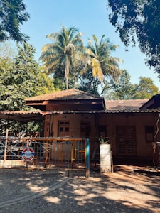 A rustic house with a tiled roof, surrounded by lush greenery and tall palm trees. The house has wooden windows and a front gate with a 'No Parking in front of gate' sign. Sunlight filters through the trees, creating dappled shadows on the ground.