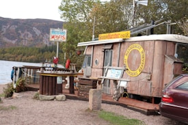 The image depicts a rustic, wooden cabin-like structure adorned with signage for 'Nessie Hunter' and 'Nessie-Serch Independent Research'. It is situated near a body of water with mountains and trees in the background. The scene includes a small outdoor seating area with a table and benches, along with a signboard displaying 'Nessie Models' and various decorations. The overall setting has an outdoor, adventurous theme.