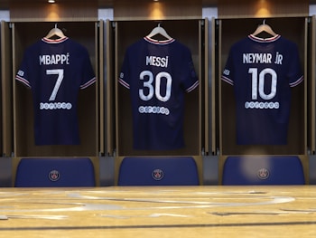 Three football jerseys displayed in a locker room setting, each with a distinct number and name on the back. The jerseys are neatly hung on wooden hangers within open lockers. The names printed above the numbers are Mbappé, Messi, and Neymar Jr, suggesting famous players. Below the jerseys, there are seats or bench areas with the PSG club logo.