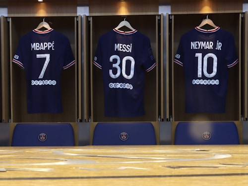 Three football jerseys displayed in a locker room setting, each with a distinct number and name on the back. The jerseys are neatly hung on wooden hangers within open lockers. The names printed above the numbers are Mbapp&eacute;, Messi, and Neymar Jr, suggesting famous players. Below the jerseys, there are seats or bench areas with the PSG club logo.