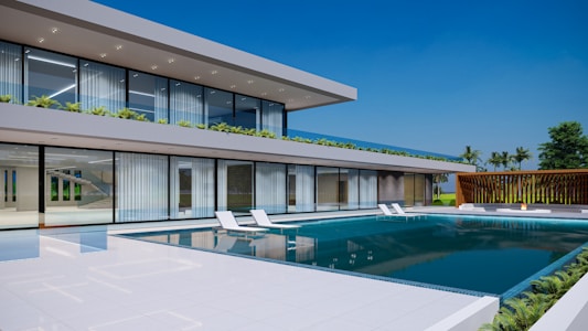 A modern two-story house with large glass windows and a sleek architectural design, highlighted by a spacious swimming pool in the foreground. The building is surrounded by greenery, including palm trees and small shrubs on the terrace. The sky is clear and blue, adding to the luxurious and serene atmosphere.