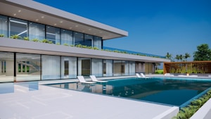 A modern two-story house with large glass windows and a sleek architectural design, highlighted by a spacious swimming pool in the foreground. The building is surrounded by greenery, including palm trees and small shrubs on the terrace. The sky is clear and blue, adding to the luxurious and serene atmosphere.