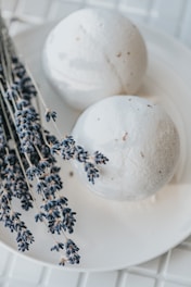 Two white bath bombs are placed on a white plate, accompanied by dried lavender sprigs. The setting is serene, with a soft focus on the lavender, highlighting its purple tones against the muted white of the bath bombs.