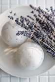 A pair of round, white bath bombs placed on a white plate, accompanied by a bunch of dried lavender sprigs. The lavenders exhibit a deep purple hue, creating a contrast with the minimalistic and clean white background and surface.