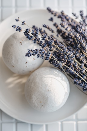 A pair of round, white bath bombs placed on a white plate, accompanied by a bunch of dried lavender sprigs. The lavenders exhibit a deep purple hue, creating a contrast with the minimalistic and clean white background and surface.