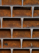 Metal grid structure with a rusty orange and brown surface, featuring rectangular openings. The grid is designed in a uniform pattern with visible aging and weathering.