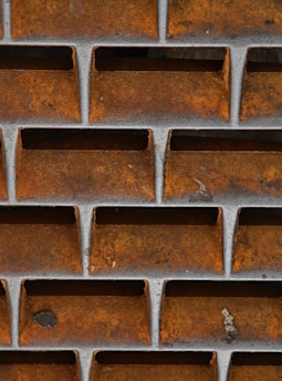 Metal grid structure with a rusty orange and brown surface, featuring rectangular openings. The grid is designed in a uniform pattern with visible aging and weathering.