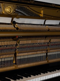 The image displays the interior mechanics of an upright piano with visible strings, hammers, and dampers. The piano's keys are partially visible at the bottom of the image. A golden metal plate frames the upper section, adding a striking contrast to the wooden components.