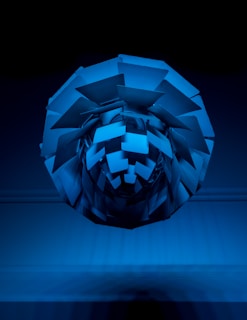 An abstract, three-dimensional geometric structure composed of various flat, angular surfaces, appearing to be suspended in mid-air against a dark background. The structure reflects light, creating varying shades of blue.