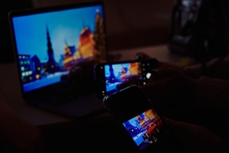 A laptop screen displaying a cityscape with illuminated buildings at night is being photographed by a smartphone. The scene captured on the laptop includes a blurred skyline with bright lights. The smartphone's screen also displays the same cityscape image, while another smartphone captures this in the foreground.