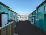 A narrow alleyway with teal-colored buildings on either side leads towards a scenic waterfront. String lights are hanging across the alley, and signage indicates a car rental service. In the background, a bay with mountains under a partly cloudy sky is visible.