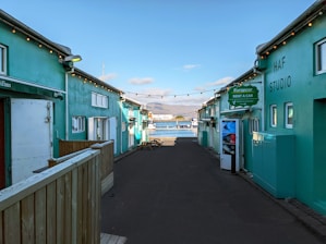 A narrow alleyway with teal-colored buildings on either side leads towards a scenic waterfront. String lights are hanging across the alley, and signage indicates a car rental service. In the background, a bay with mountains under a partly cloudy sky is visible.