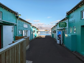 A narrow alleyway with teal-colored buildings on either side leads towards a scenic waterfront. String lights are hanging across the alley, and signage indicates a car rental service. In the background, a bay with mountains under a partly cloudy sky is visible.
