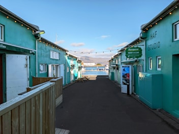 A narrow alleyway with teal-colored buildings on either side leads towards a scenic waterfront. String lights are hanging across the alley, and signage indicates a car rental service. In the background, a bay with mountains under a partly cloudy sky is visible.