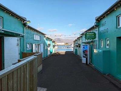 A narrow alleyway with teal-colored buildings on either side leads towards a scenic waterfront. String lights are hanging across the alley, and signage indicates a car rental service. In the background, a bay with mountains under a partly cloudy sky is visible.
