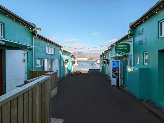 A narrow alleyway with teal-colored buildings on either side leads towards a scenic waterfront. String lights are hanging across the alley, and signage indicates a car rental service. In the background, a bay with mountains under a partly cloudy sky is visible.