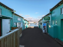 A narrow alleyway with teal-colored buildings on either side leads towards a scenic waterfront. String lights are hanging across the alley, and signage indicates a car rental service. In the background, a bay with mountains under a partly cloudy sky is visible.