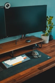 A modern, organized desk setup featuring a widescreen monitor with a webcam on top, a mechanical keyboard with colorful keys, a wireless mouse, and a plant in a white pot. The desk surface is wooden with a black mat underneath the keyboard and mouse, creating a neat and functional workspace.