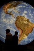 A large illuminated globe showing a detailed view of North and South America. Two silhouetted figures stand in the foreground, facing the globe. The scene takes place in a dimly lit setting, emphasizing the vibrant details and colors of the Earth depicted on the globe.