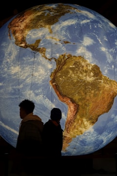 A large illuminated globe showing a detailed view of North and South America. Two silhouetted figures stand in the foreground, facing the globe. The scene takes place in a dimly lit setting, emphasizing the vibrant details and colors of the Earth depicted on the globe.