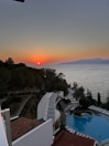A picturesque sunset over a calm sea, with the orange sun setting on the horizon. Below, a winding path leads down a hillside covered in trees, while a swimming pool is nestled among palm trees and sun loungers. The scene captures both natural beauty and resort-like comforts.