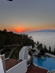A picturesque sunset over a calm sea, with the orange sun setting on the horizon. Below, a winding path leads down a hillside covered in trees, while a swimming pool is nestled among palm trees and sun loungers. The scene captures both natural beauty and resort-like comforts.